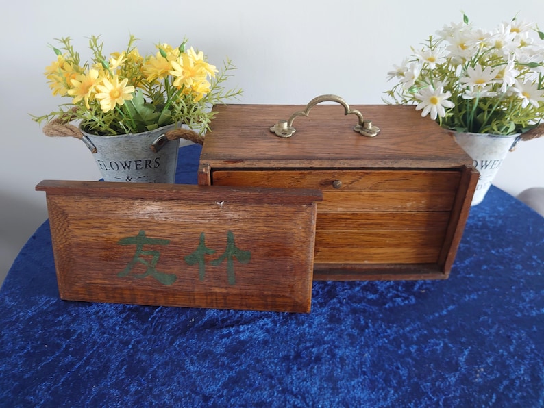 May include: A wooden box with a brass handle and a smaller wooden box in front of it. The smaller box has green text on it. Two small metal buckets with flowers are on either side of the boxes.