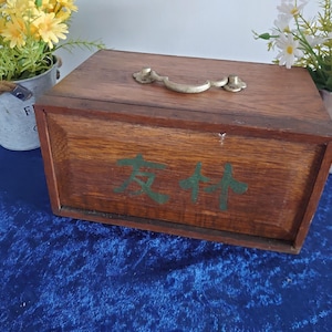 May include: A rectangular wooden box with a brass handle and hinges. The box is stained a dark brown color and has green Chinese characters painted on the front. The box is sitting on a blue surface.