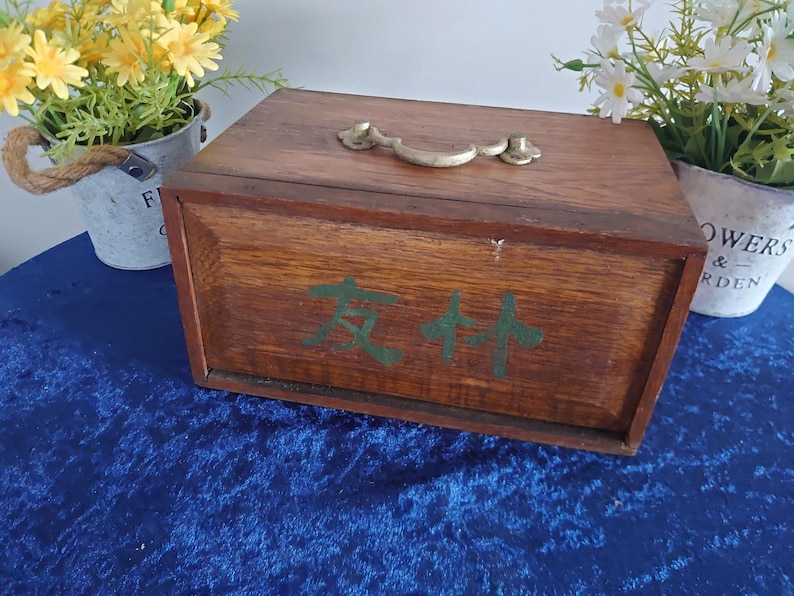 May include: A rectangular wooden box with a brass handle and hinges. The box is stained a dark brown color and has green Chinese characters painted on the front. The box is sitting on a blue surface.