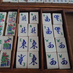 May include: A vintage Mahjong set with ivory-colored tiles arranged in a wooden box. The tiles feature Chinese characters and colorful illustrations of flowers and plants. The set includes dice and is ready for play.