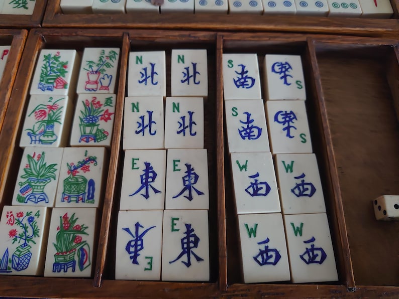 May include: A vintage Mahjong set with ivory-colored tiles arranged in a wooden box. The tiles feature Chinese characters and colorful illustrations of flowers and plants. The set includes dice and is ready for play.