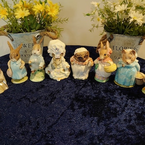 May include: A collection of ceramic figurines featuring various animal characters, including rabbits, bears, and a duck, dressed in detailed outfits. The figurines are arranged on a dark blue surface, with two metal buckets of flowers in the background.