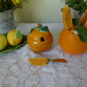 Può includere: Un set di stoviglie in ceramica a tema arancia, che comprende una zuccheriera con coperchio, due piccoli contenitori a forma di limone, un grande contenitore a forma di arancia con fette di arancia e un piccolo coltello da burro a forma di fetta di arancia. Gli articoli sono su una tovaglia bianca con un motivo a pizzo.