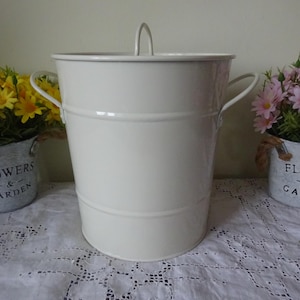 May include: White metal storage bin with a lid and two handles. The bin is sitting on a white lace tablecloth. The text "Flowers & Garden" is printed on a gray metal planter in the background.