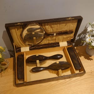 May include: An open antique vanity set with a brown faux tortoiseshell finish. The set includes a hand mirror, two brushes, and a shoe horn. The interior is lined with a cream-colored fabric. The set is displayed on a wooden surface.