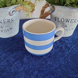 Puede incluir: Una taza de cerámica con rayas horizontales azules y blancas. La taza tiene un interior blanco y un asa curva. Dos pequeños cubos de metal con las palabras "FLOWERS & GARDEN" están en el fondo.
