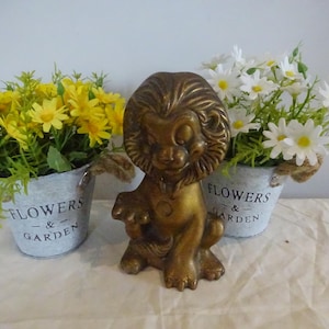 May include: A golden lion figurine sits between two small metal buckets of flowers. One bucket holds yellow daisies, the other white daisies. The buckets have rope handles and the words "FLOWERS & GARDEN" printed on them.