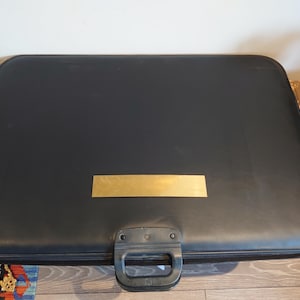 Puede incluir: Un maletín negro rectangular con una placa rectangular dorada en la parte delantera. El maletín tiene un asa y esquinas redondeadas. El maletín está sobre una superficie de madera.