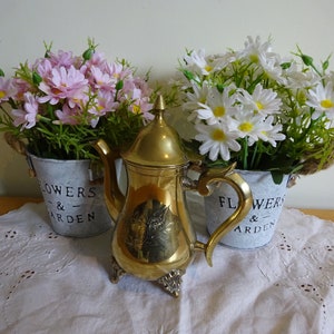 May include: A gold metal teapot with a decorative design sits on a white lace tablecloth between two white metal flower pots with the text "Flowers & Garden" on them. The pots are filled with pink and white artificial flowers.