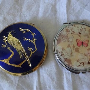Op de afbeelding: Twee vintage compact spiegels. Eén heeft een blauwe achtergrond met een gouden fazant en takken. De andere heeft een bloemenpatroon met een roze vlinder.