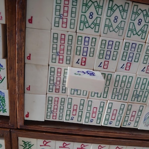 May include: A close-up of a vintage Mahjong set. The ivory-colored tiles are arranged in a wooden box, displaying various symbols and numbers in red, green, and blue. Some tiles feature floral designs. The set is a classic game.