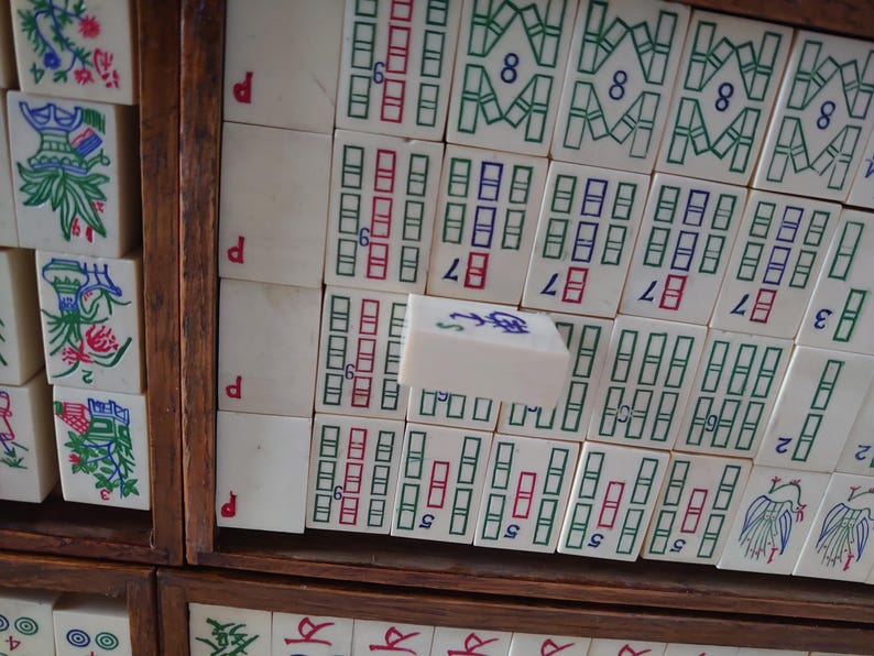 May include: A close-up of a vintage Mahjong set. The ivory-colored tiles are arranged in a wooden box, displaying various symbols and numbers in red, green, and blue. Some tiles feature floral designs. The set is a classic game.