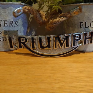 Puede incluir: Un emblema Triumph plateado y negro, con la palabra "TRIUMPH" en letras negras en negrita, se muestra sobre una superficie de madera. El emblema es curvo y tiene un borde negro. Dos macetas de metal galvanizado con asas de cuerda est&aacute;n en el fondo.