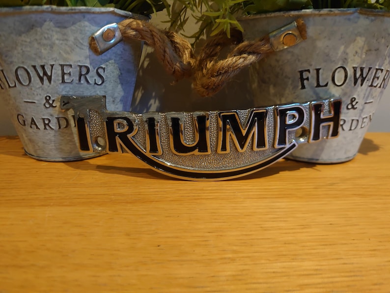 Puede incluir: Un emblema Triumph plateado y negro, con la palabra "TRIUMPH" en letras negras en negrita, se muestra sobre una superficie de madera. El emblema es curvo y tiene un borde negro. Dos macetas de metal galvanizado con asas de cuerda est&aacute;n en el fondo.