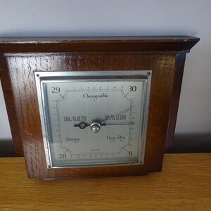 May include: A vintage wooden barometer with a silver-colored face. The barometer has a rectangular shape with a dark brown wooden frame. The face displays the words "Rain", "Fair", and "Changeable". The barometer is designed to measure atmospheric pressure.