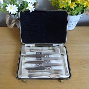 May include: A vintage cutlery set in an open case. The case has a black interior and contains knives and forks with white, possibly mother-of-pearl, handles. The case is on a wooden surface.
