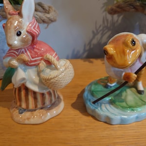 Può includere: Due statuine in ceramica: un coniglio con giacca rossa e una rana con canna da pesca. Il coniglio tiene un cestino e indossa un cappello. La rana è su una ninfea. Le statuine sono dipinte con dettagli.