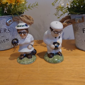 Può includere: Un paio di saliere e pepiere in ceramica raffiguranti personaggi del bowling. Le figure indossano occhiali e cappelli, tenendo in mano delle palle da bowling. Le saliere sono su una superficie di legno, con vasi di fiori sullo sfondo.