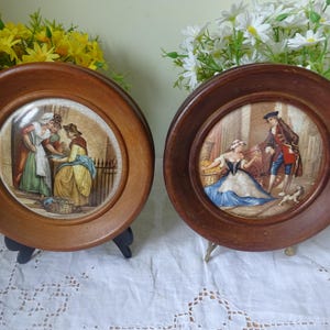 Puede incluir: Dos platos decorativos antiguos con marcos de madera. Cada plato presenta una escena detallada y colorida de personas con trajes de época. Los platos se exhiben sobre pequeños soportes, lo que sugiere que están destinados a la exhibición. Los marcos son de color marrón cálido.