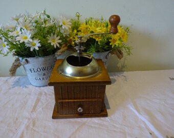 May include: A wooden coffee grinder with a metal top and a crank handle. The grinder has a drawer on the bottom and is sitting on a white tablecloth. There are two flower pots with white daisies and yellow flowers in the background.