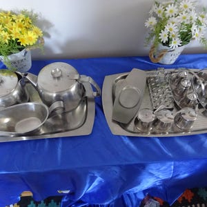 Puede incluir: Dos juegos de servir de acero inoxidable sobre un mantel azul. Uno incluye una tetera, una crema y un azucarero. El otro tiene un rallador, recipientes para condimentos y una bandeja. Flores amarillas y blancas en macetas decorativas están en el fondo.