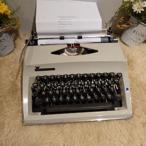 Op de afbeelding: Een vintage, crèmekleurige typemachine met zwarte toetsen en een ingevoegd vel papier. De typemachine heeft een chromen afwerking en een rode lintindicator. De afbeelding is van bovenaf genomen, met bloemen in kleine metalen emmers op de achtergrond.