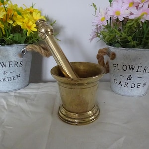 Könnte beinhalten: Ein Messing-Mörser und -Stößel-Set. Der Mörser hat eine zylindrische Schüssel und der Stößel ist lang. Der Mörser steht auf einer weißen Oberfläche, im Hintergrund zwei kleine Metalleimer mit Blumen. Die Eimer haben Seilgriffe und die Aufschrift "FLOWERS & GARDEN".