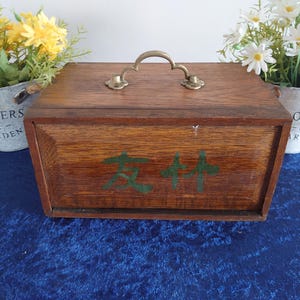 May include: A wooden box with a brass handle and green Chinese characters on the front. The box is brown and sits on a blue surface. Two small metal buckets with flowers are on either side of the box.