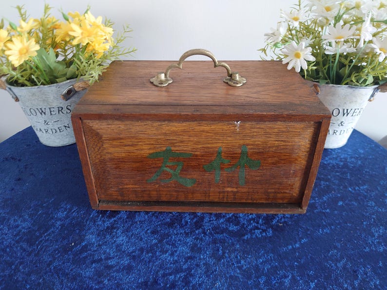 May include: A wooden box with a brass handle and green Chinese characters on the front. The box is brown and sits on a blue surface. Two small metal buckets with flowers are on either side of the box.