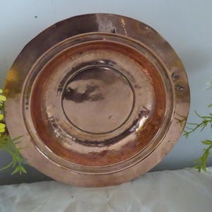 Peut inclure: Une assiette ronde de couleur cuivre avec une zone centrale en retrait et des anneaux concentriques. L'assiette a une surface réfléchissante et semble être en métal. L'assiette est probablement utilisée pour servir ou exposer.