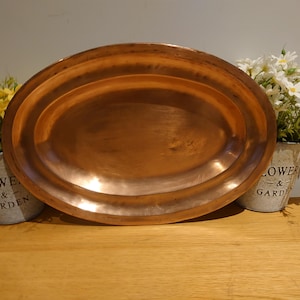 Peut inclure: Un grand plateau ovale en cuivre avec une finition polie. Le plateau est présenté sur une surface en bois, avec de petits seaux en métal de fleurs de chaque côté. Les seaux portent les mots "Flower & Garden".