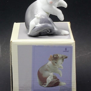 Puede incluir: Figura de porcelana de un gato y un ratón. El gato es blanco y gris, con detalles rosas, y está sentado con el ratón a su lado. La figura está sobre una caja con una imagen de la figura y el texto "05236 GATITO PASMADO CAT AND MOUSE".