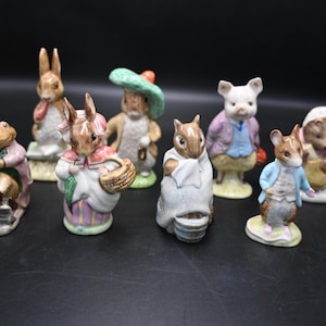 May include: A group of ceramic animal figurines, including rabbits, a frog, and a squirrel. The figures are dressed in detailed clothing and painted in pastel colours. The collection is set against a dark background.