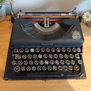 May include: A vintage black Imperial Model 7 typewriter with a QWERTY keyboard. The keys have white lettering. The typewriter features a silver carriage and the words "Imperial" and "The Good Companion". It sits on a wooden surface.