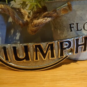 Puede incluir: Un emblema Triumph cromado con letras negras, frente a un cubo de metal galvanizado borroso. El cubo tiene las palabras "FLOWERS & GARDEN". El emblema est&aacute; sobre una superficie de madera.