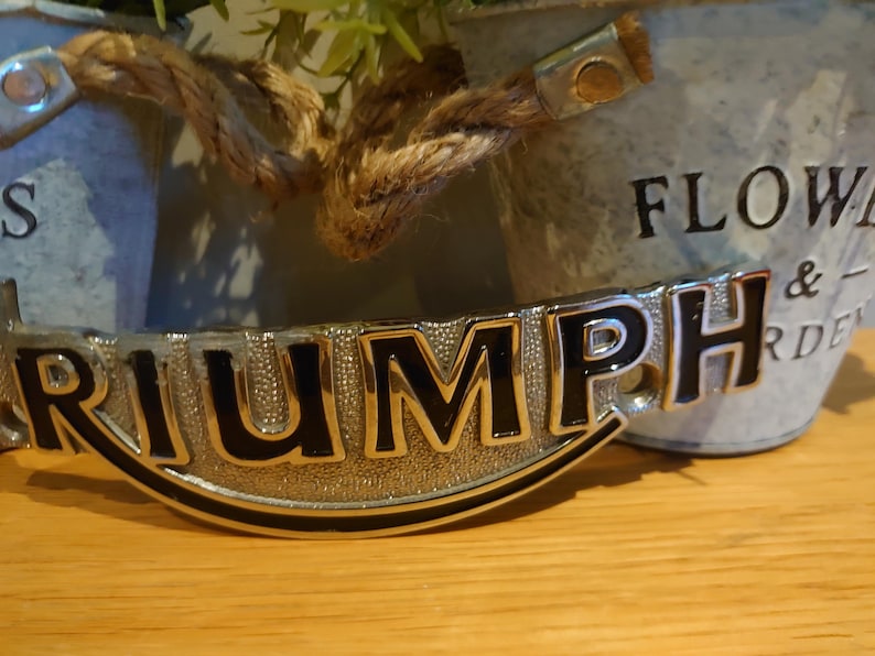 Puede incluir: Un emblema Triumph cromado con letras negras, frente a un cubo de metal galvanizado borroso. El cubo tiene las palabras "FLOWERS & GARDEN". El emblema est&aacute; sobre una superficie de madera.