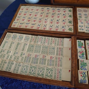 May include: A vintage Mahjong set with ivory-colored tiles arranged in wooden trays. The tiles feature Chinese characters and symbols in red, green, and blue. The set is displayed on a blue surface.