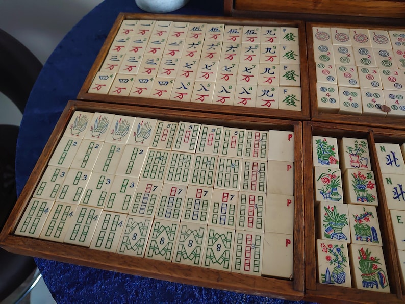 May include: A vintage Mahjong set with ivory-colored tiles arranged in wooden trays. The tiles feature Chinese characters and symbols in red, green, and blue. The set is displayed on a blue surface.