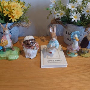 Puede incluir: Colección de figuras de cerámica con personajes de los cuentos de Beatrix Potter. Las figuras incluyen conejos, un erizo, un pato y un cerdo, todos con trajes coloridos y detallados. También hay un pequeño libro con el título "Beatrix Potter".