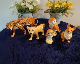 Op de afbeelding: Een verzameling keramische dierenbeeldjes, waaronder een tijger met oranje en zwarte strepen, een bruine cheetah, twee uilen en een corgi. De beeldjes staan op een donkerblauwe ondergrond, met kleine metalen emmers met bloemen op de achtergrond.