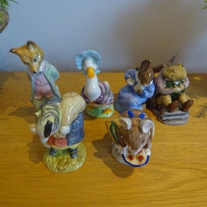 Może przedstawiać: Kolekcja sześciu ceramicznych figurek przedstawiających postacie z bajki dla dzieci. Figurki obejmują lisa, gęś, królika, mysz i borsuka, wszystkie w kolorowych strojach. Figurki są wystawione na drewnianej powierzchni.