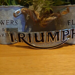 Puede incluir: Un emblema Triumph plateado con letras negras, colocado sobre una superficie de madera. El emblema es curvo y tiene una superficie texturizada. Dos macetas de metal galvanizado con asas de cuerda y vegetaci&oacute;n en el fondo.