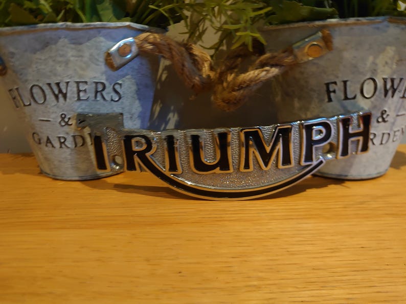 Puede incluir: Un emblema Triumph plateado con letras negras, colocado sobre una superficie de madera. El emblema es curvo y tiene una superficie texturizada. Dos macetas de metal galvanizado con asas de cuerda y vegetaci&oacute;n en el fondo.