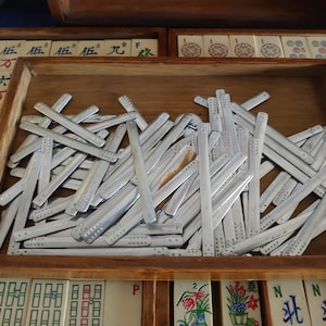 May include: A wooden tray filled with silver-colored Mahjong scoring sticks. The sticks are rectangular with rounded ends and small indentations. The tray sits among other Mahjong tiles and trays.