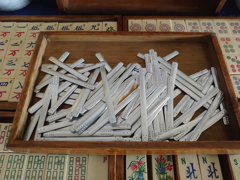 May include: A wooden tray filled with silver-colored Mahjong scoring sticks. The sticks are rectangular with rounded ends and small indentations. The tray sits among other Mahjong tiles and trays.