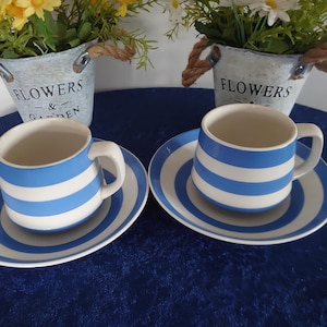 May include: Two matching blue and white striped teacups and saucers are displayed on a dark blue surface. The cups and saucers have a classic design. Two small galvanized metal buckets with flowers are in the background.