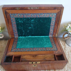 Puede incluir: Caja de escritura de madera abierta, con interior de terciopelo verde y borde decorativo. La caja tiene pequeños compartimentos y un cierre de latón. El exterior es de color marrón cálido. La caja está abierta.