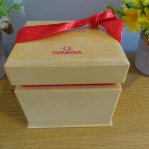 Op de afbeelding: Een lichtbeige Omega horlogedoos met een rood lint erbovenop. Het Omega-logo is rood op het deksel gedrukt. De doos staat op een houten oppervlak, met bloemen op de achtergrond.