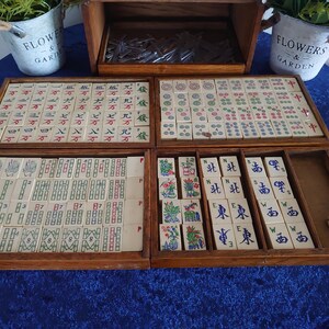 May include: A vintage wooden Mahjong set with tiles featuring Chinese characters and symbols. The set includes multiple trays and a storage box, with dice and decorative elements. The tiles are cream-colored with red, green, and blue markings.
