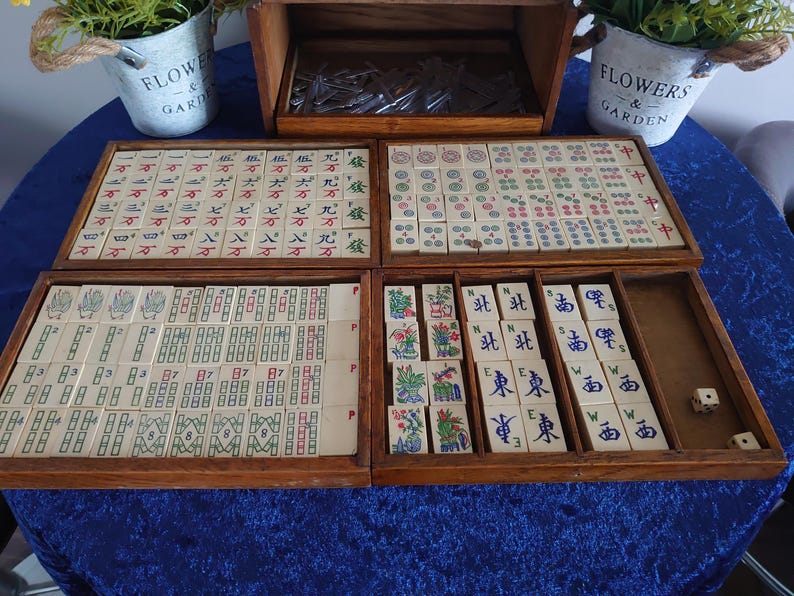 May include: A vintage wooden Mahjong set with tiles featuring Chinese characters and symbols. The set includes multiple trays and a storage box, with dice and decorative elements. The tiles are cream-colored with red, green, and blue markings.
